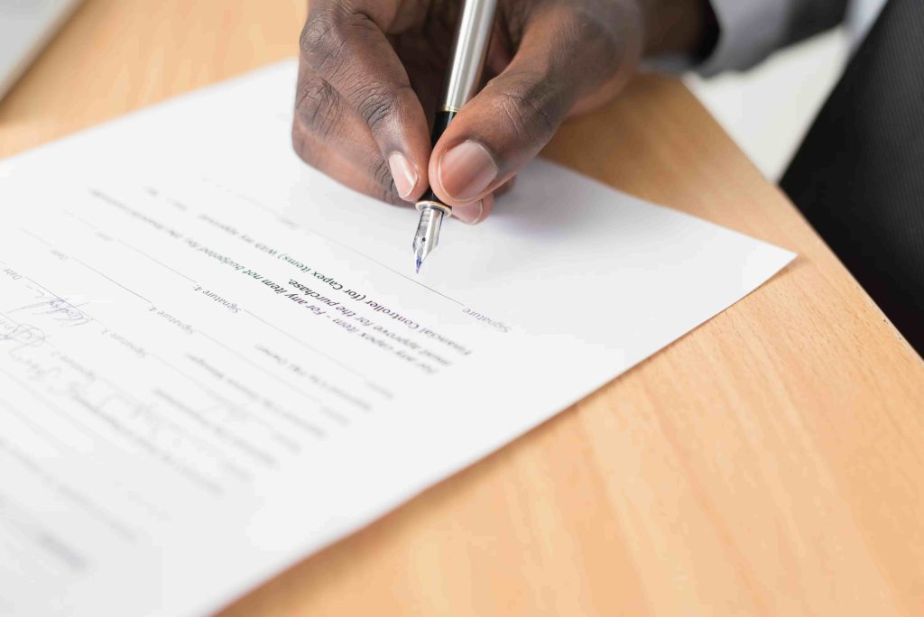 Eine Person unterschreibt einen Vertrag auf Papier an einem Tisch, welcher von einem Anwalt für Gesellschaftsrecht geprüft wurde.